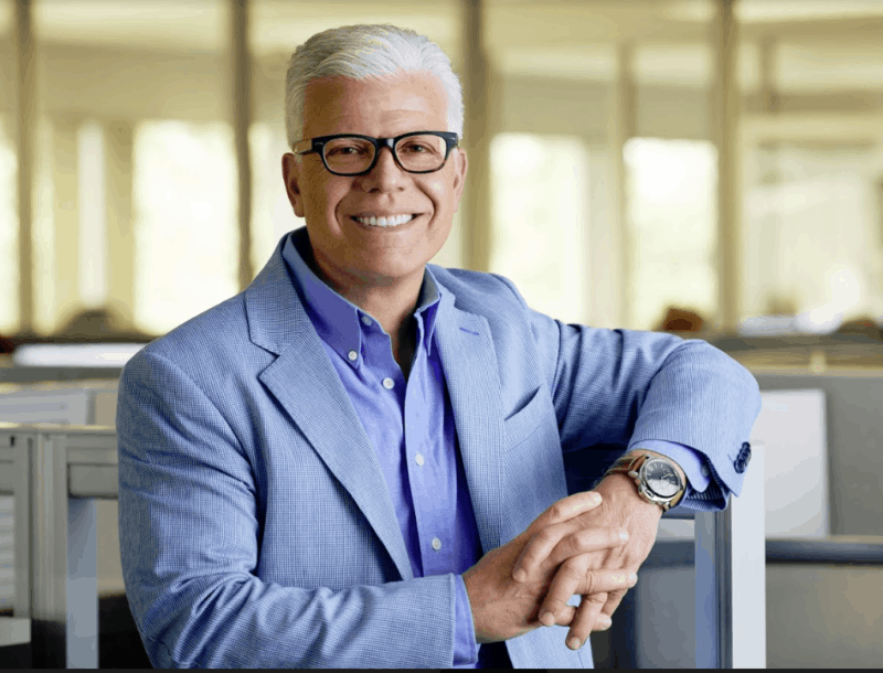 Vince Passione headshot in an office setting. Vince smiles, wears a suit and button-up shirt with white hair and eye glasses.